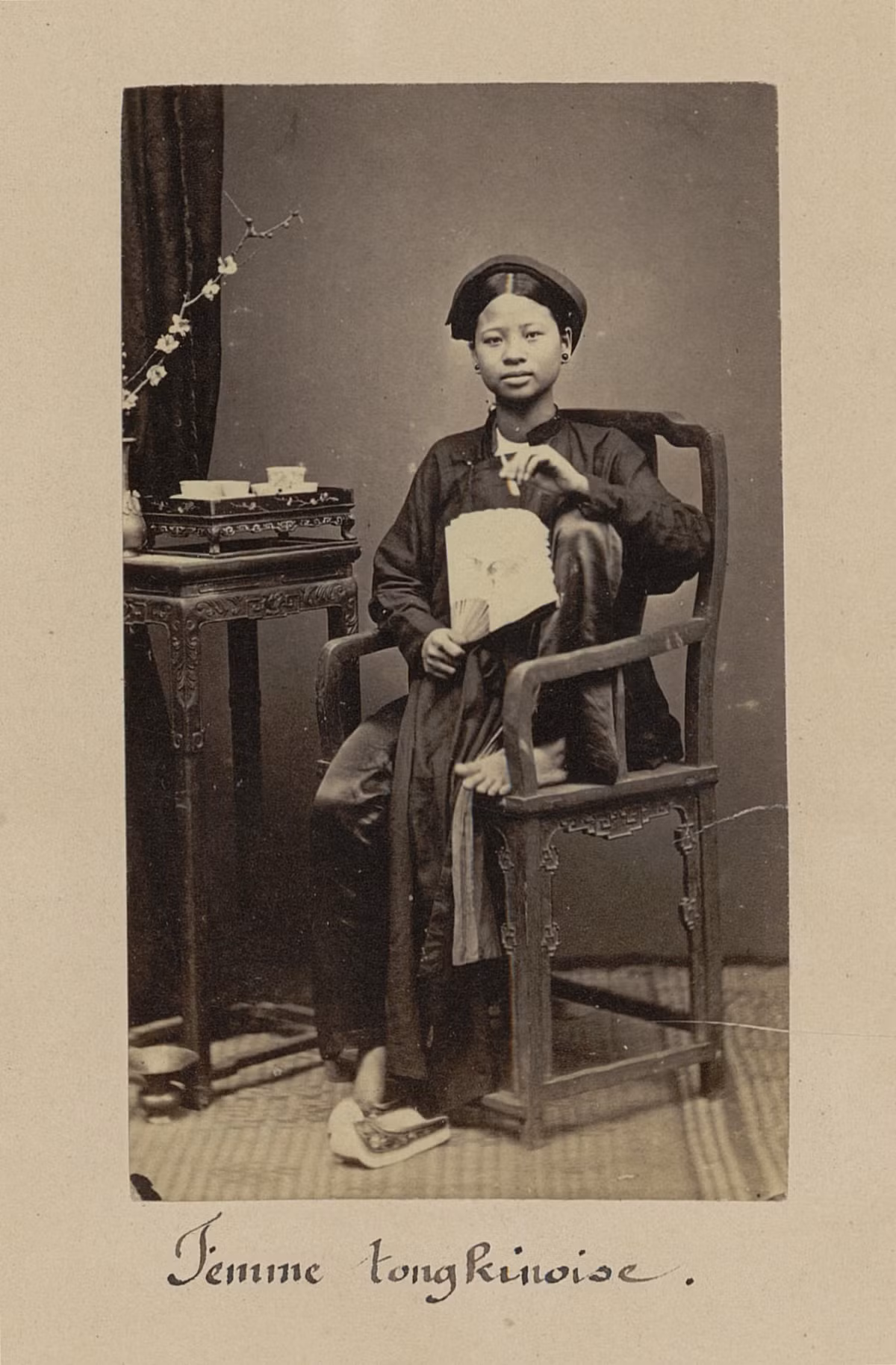 Chân dung thiếu nữ xứ Bắc Kỳ mặc trang phục truyền thống, tay cầm quạt và ngồi trên ghế gỗ.