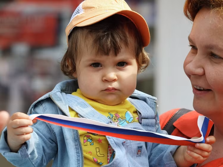 Một phụ bế bé gái cầm tuy băng với ba màu của quốc kỳ Nga trong lễ kỷ niệm ở Kazan.