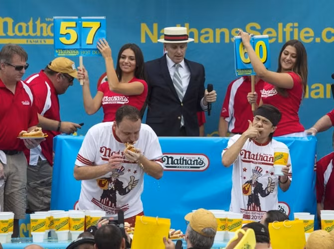 Joey Chestnut (trái) and Matt Stonie (phải) thi ăn nhanh hot dog. Vào lúc gần kết thúc cuộc thi, Matt Stonie dẫn trước Joey Chestnut với tỷ số 60-57.