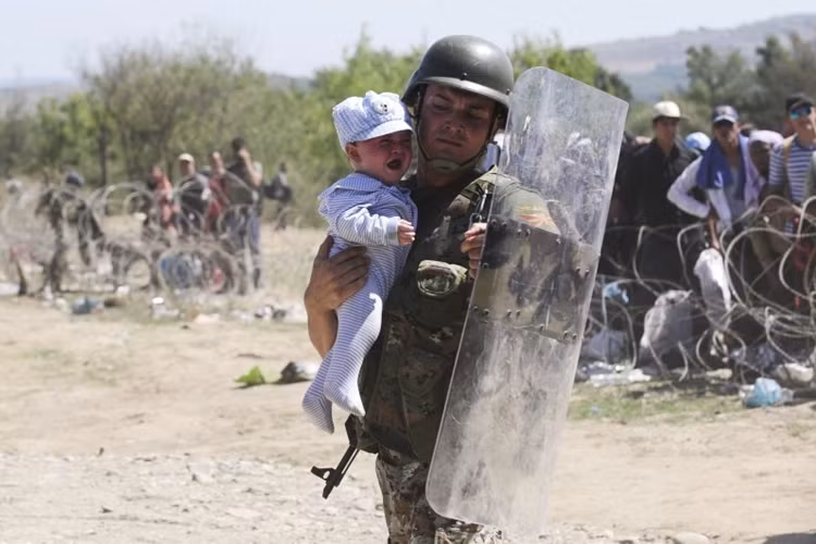 Đối với nhiều người tị nạn, Macedonia chỉ là nước quá cảnh để họ tràn vào Serbia và sau đó vào Hungary, trên đường sang Tây Âu. Trong ảnh: Cảnh sát Macedonia bế một bé tị nạn vượt biên.