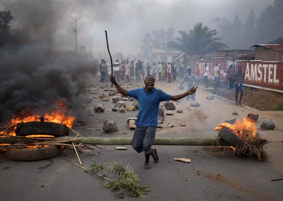 Hiến pháp cũng như Thỏa thuận hòa bình và hòa giải Arusha quy định tổng thống Burundi không được tại nhiệm quá hai nhiệm kỳ liên tiếp. Trong ảnh: Một người biểu tình vung gậy trước "chiến lũy" được tạo bằng cây và lốp xe đốt cháy.