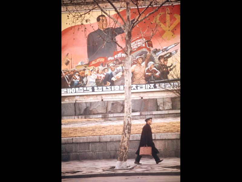 Một người đàn ông bước qua bức tranh tường có hình ảnh lãnh tụ Kim Nhật Thành và lực lượng vũ trang Triều Tiên.