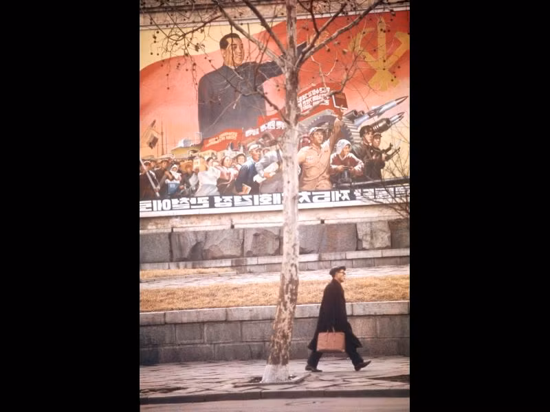 Một người đàn ông bước qua bức tranh tường có hình ảnh lãnh tụ Kim Nhật Thành và lực lượng vũ trang Triều Tiên.