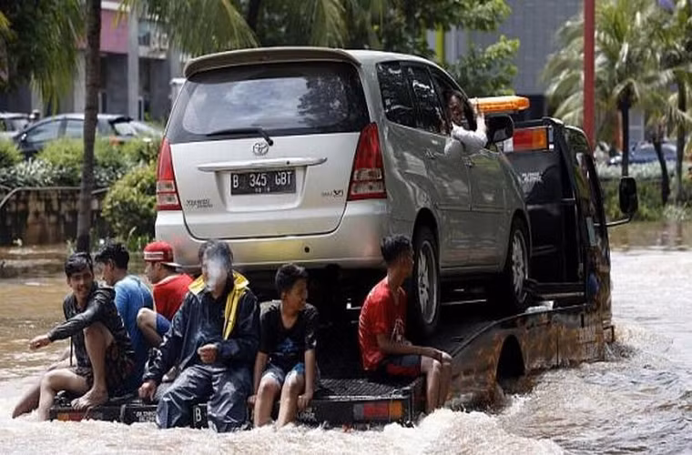 1 số người ngồi sau chiếc xe tải đang chuyển 1 chiếc ô tô khỏi khu vực ngập lụt.