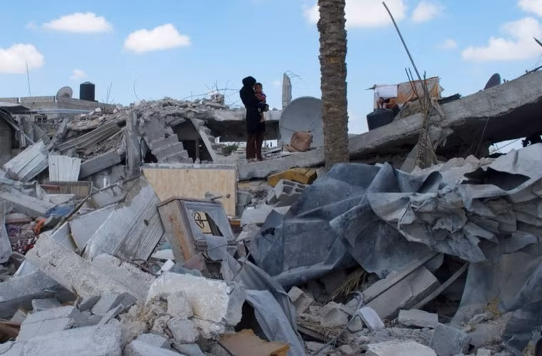 Người phụ nữ ôm đứa trẻ đứng trên đống đổ nát của căn nhà ở Khuzaa, khu vực này cũng đã bị oanh tạc khủng khiếp.