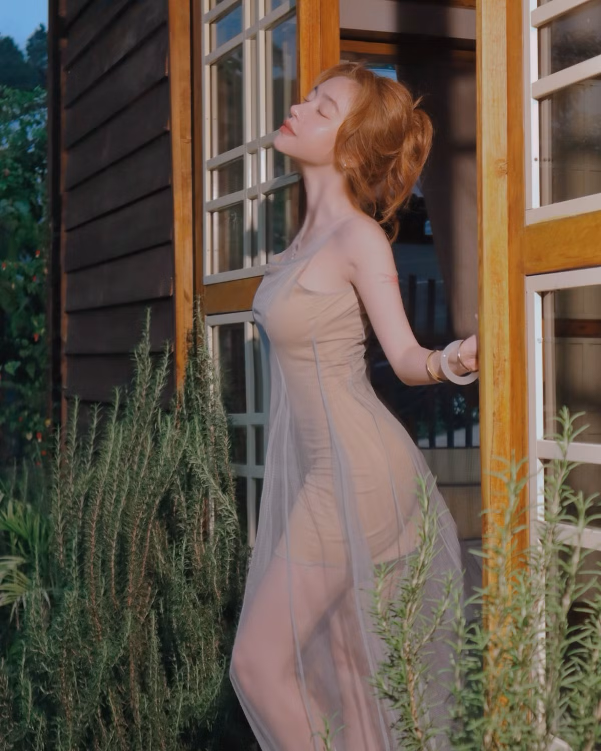 Đứng nghiêng bên khung cửa gỗ, đón ánh hoàng hôn nhẹ buông, Elly Trần khoe trọn thần thái kiêu sa, gợi cảm nhưng vẫn phảng phất nét mơ màng, dịu dàng. Lối trang điểm trong trẻo và kiểu tóc búi nhẹ nhàng càng làm nổi bật vẻ đẹp trong trẻo không tuổi của “bà mẹ hai con”.