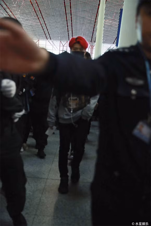 Tại sân bay, ngôi sao nam ăn mặc trẻ trung, sành điệu với quần jeans, áo nỉ hoạ tiết và mái tóc đỏ. Anh đeo khẩu trang, giữ thái độ lạnh lùng và còn được hơn 10 vệ sĩ đi theo cùng để đảm bảo an ninh.