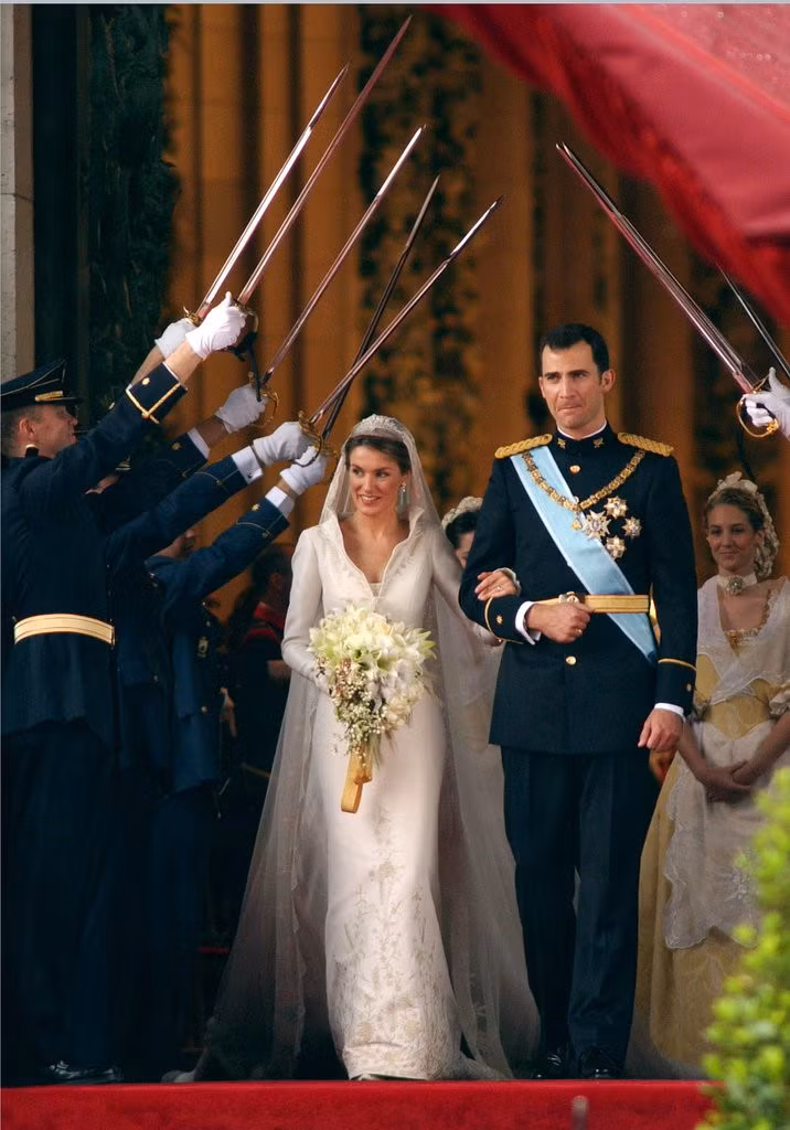 Hoàng hậu Letizia Ortiz trong lễ cưới với nhà vua Felipe VI của Tây Ban Nha và năm 2004. Váy cưới của bà được thiết kế bởi Manuel Pertegaz với kiểu dáng cổ lật tôn lên vẻ quyền lực và uy quyền của một bà hoàng.