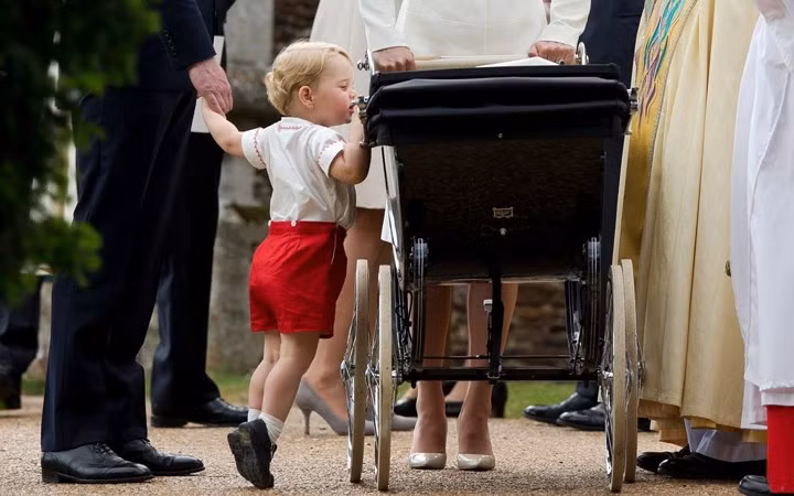 Tiểu Hoàng tử George kiễng chân nhìn em gái Charlotte nằm trong xe nôi trong lễ đặt tên thánh cho tiểu Công chúa ở Sandringham, Norfolk.