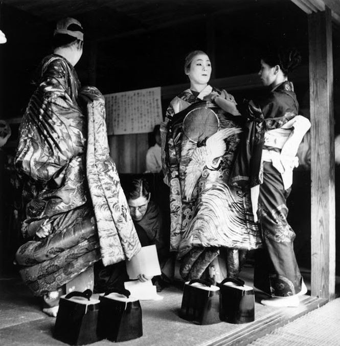 Những geisha đang chỉnh trang trước khi biểu diễn, gương mặt được tô vẽ trắng bệch đặc trưng nhưng chưa đội tóc giả. Trang phục để biểu diễn trông đồ sộ hơn nhiều so với những bộ kimono thường thấy, đặc biệt là đôi dép cao cả gang tay. Ảnh: Three Lions