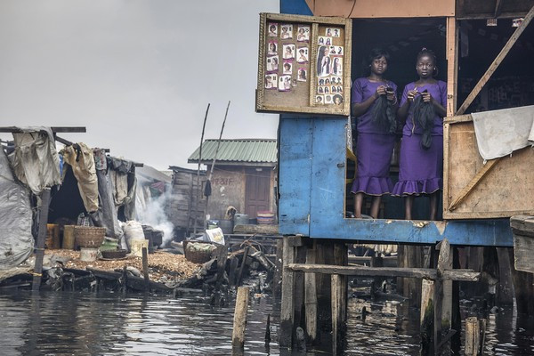 Hình ảnh hai người phụ nữ tại một cửa tiệm làm tóc tại khu ổ chuột ở Makoto, Nigeria. Trước đây, dòng sông này là nơi mà những người dân nơi đây ra tắm giặt, sinh hoạt. Nhưng giờ đây, nước sông đã bị ô nhiễm nặng nề với một màu đen xì, rác nổi...