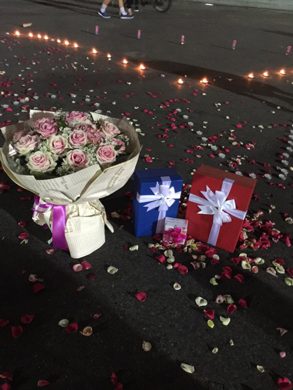 Cùng với dòng chữ "LOVE", ở chính giữa trái tim lớn được xếp bằng nến có đặt một bó hoa hồng và 3 hộp quả từ lớn, nhỏ. Đây là những món quà mà chàng trai trẻ tên Hoàng Anh - chủ nhân của màn cầu hôn này dành tặng cho bạn gái của mình.