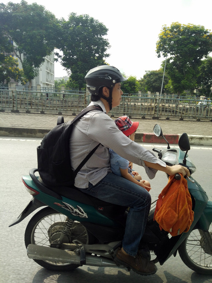 Một ông bố điều khiển xe máy bằng một tay, tay còn lại dùng ôm con di chuyển trên đường Nguyễn Cơ Thạch (Nam Từ Liêm, Hà Nội).