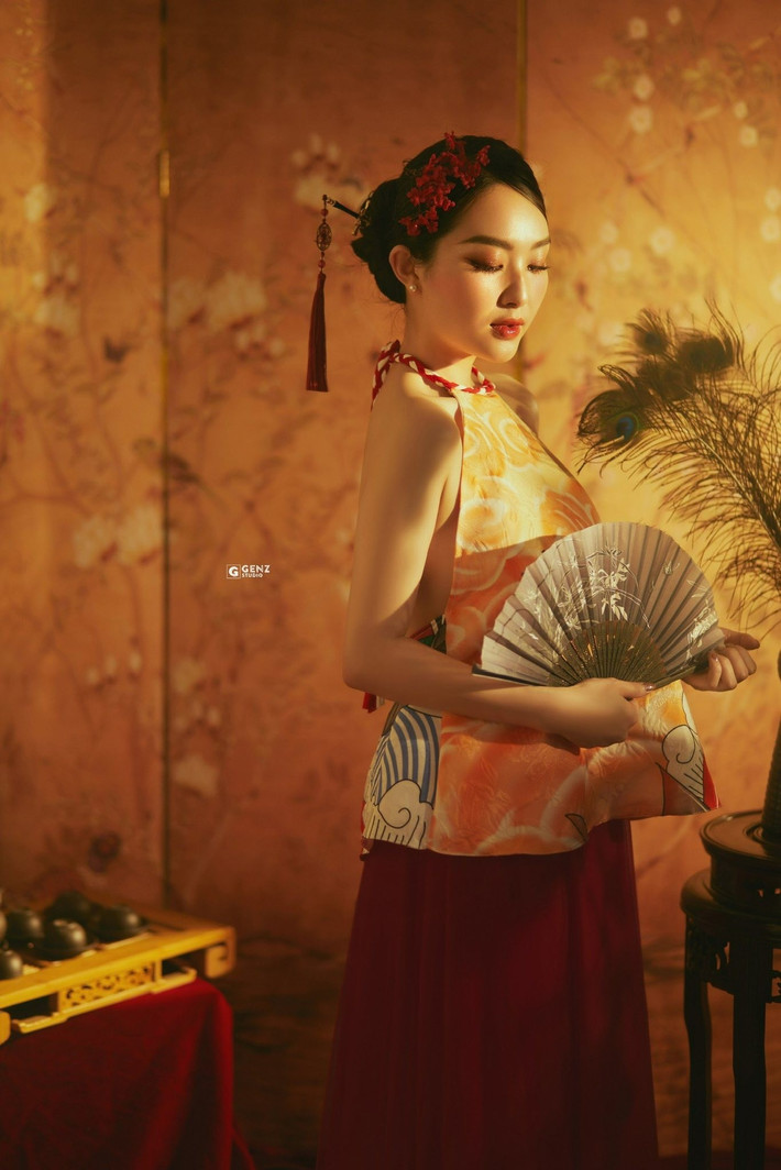 Cô sử dụng quạt giấy và hoa đỏ cài tóc làm điểm nhấn, tạo nên thần thái của một nàng thơ hoài cổ giữa không gian bài trí đậm chất cung đình. (Ảnh: FBNV)