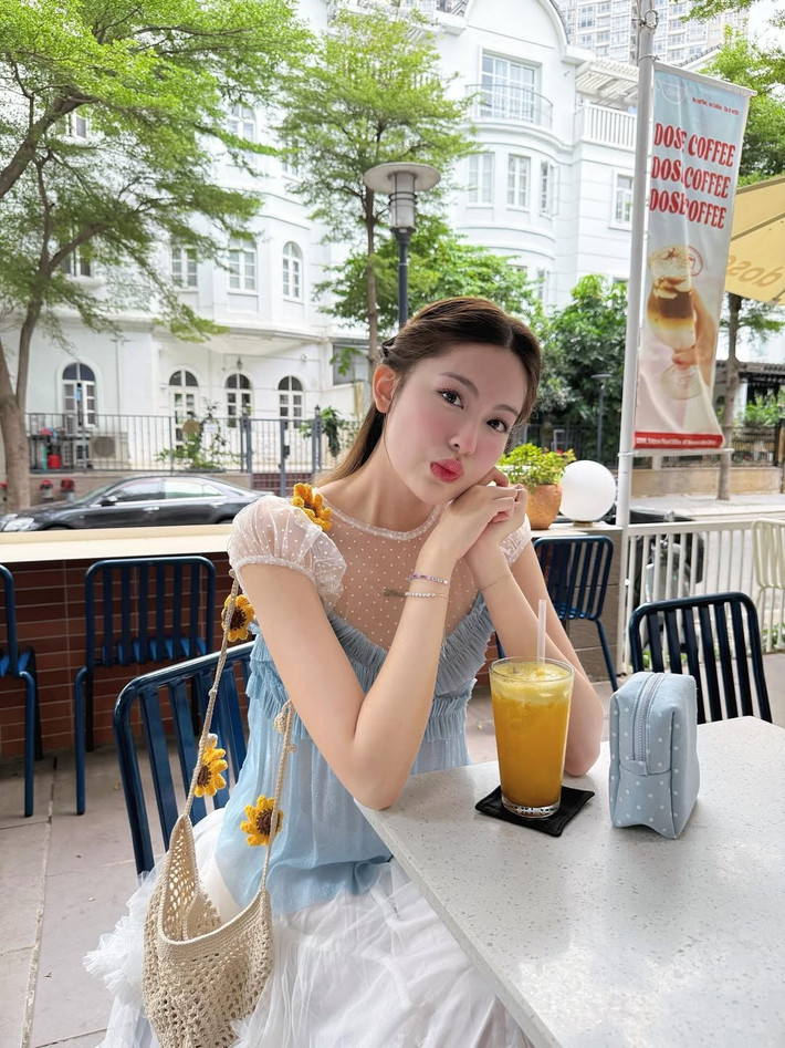 Điểm nhấn của trang phục là chi tiết bèo nhún tinh tế cùng những bông hoa nhỏ xinh được cài trên vai và túi xách, càng làm tăng thêm vẻ đáng yêu và dịu dàng.