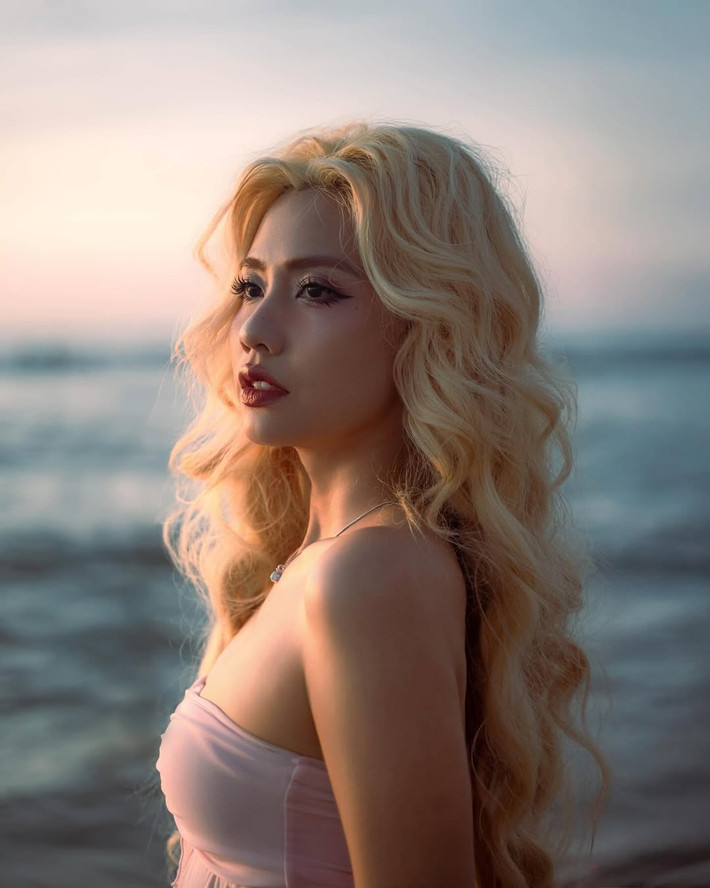 Cô nàng khoác lên mình bộ bikini màu hồng pastel dịu nhẹ, với phần áo quây xếp nhún bồng bềnh và quần cạp cao đồng điệu. Mái tóc bạch kim quen thuộc được uốn xoăn bồng bềnh, buông xõa tự nhiên, càng tôn lên vẻ đẹp mềm mại và nữ tính của cô.