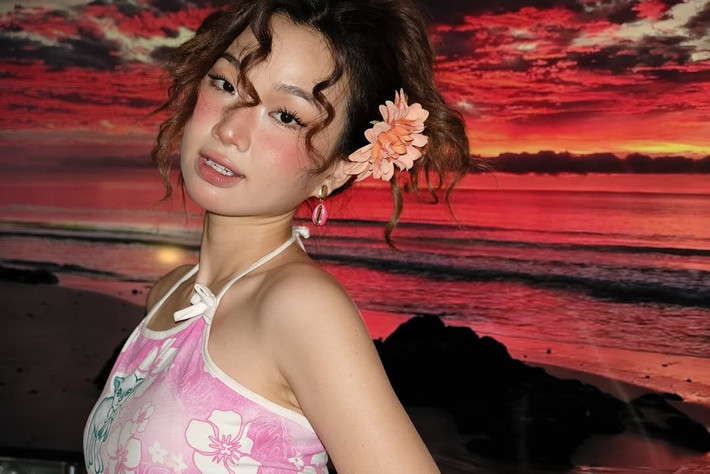 Mái tóc xoăn tít, búi cao hai bên hoặc tạo kiểu messy bun, mang đến vẻ nghịch ngợm, đáng yêu và đầy sức sống. Bông hoa màu cam rực rỡ cài trên tóc là điểm nhấn nổi bật, hài hòa với màu sắc của trang phục. Cô nàng cũng đeo vòng tay chunky màu hồng trong suốt, càng tăng thêm vẻ trẻ trung, phóng khoáng.