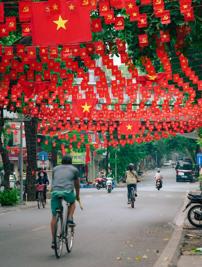 Trong không gian tràn ngập màu cờ, cuộc sống thường nhật của người dân Thủ đô vẫn diễn ra nhịp nhàng. Những người đi xe đạp, xe máy, hay những gánh hàng rong vẫn lặng lẽ di chuyển dưới những dải cờ. (Ảnh: Blog Của Rọt)