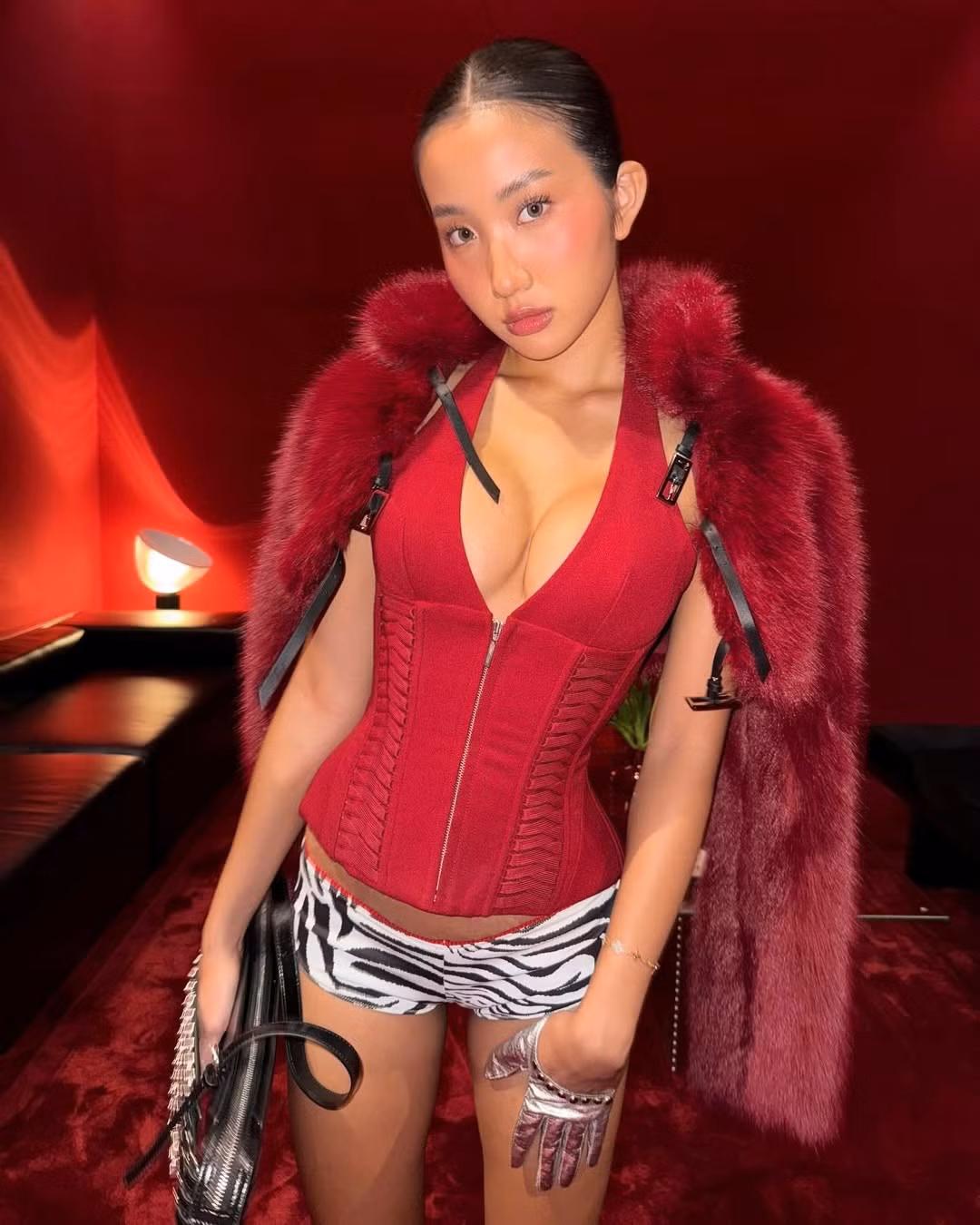 Với outfit đầu tiên, cô nàng diện áo corset màu đỏ rực rỡ với phần cổ yếm xẻ sâu, khoe trọn vòng một gợi cảm và bờ vai thon thả. Chiếc áo được phối cùng quần shorts họa tiết da ngựa vằn, tạo sự tương phản thú vị và tăng thêm vẻ cá tính.