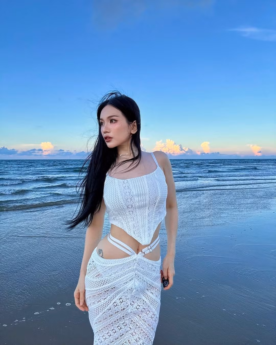 Tông trắng chủ đạo hòa quyện cùng khung cảnh biển xanh cát trắng giúp tổng thể trở nên mơ màng, vừa ngọt ngào vừa mạnh mẽ. Dưới ánh sáng hoàng hôn, từng đường nét trên bộ váy càng thêm nổi bật, tạo hiệu ứng thị giác đầy mê hoặc.