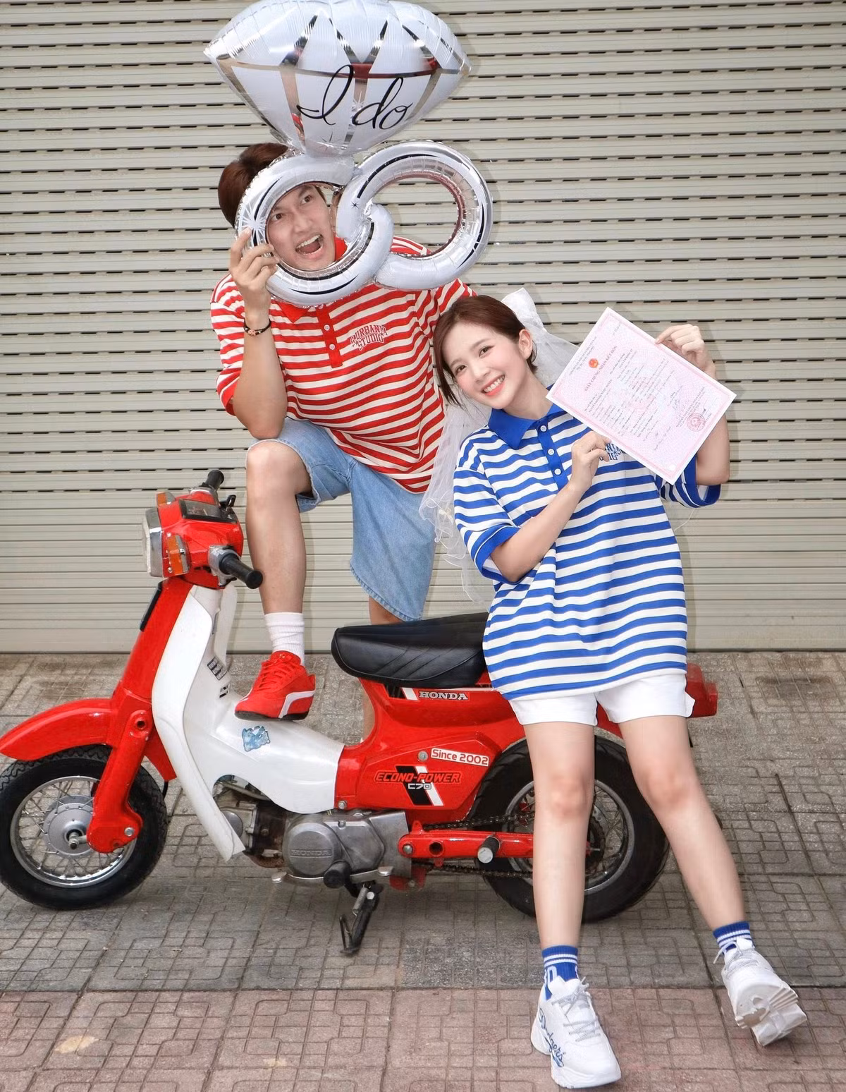 Ngọc Huyền duyên dáng ngồi trên chiếc xe máy cũ màu đỏ, trong khi Đình Tú đứng cạnh, tay cầm chiếc bóng bay độc đáo. Cả hai đều diện trang phục áo polo kẻ sọc năng động, toát lên vẻ hạnh phúc và mãn nguyện.