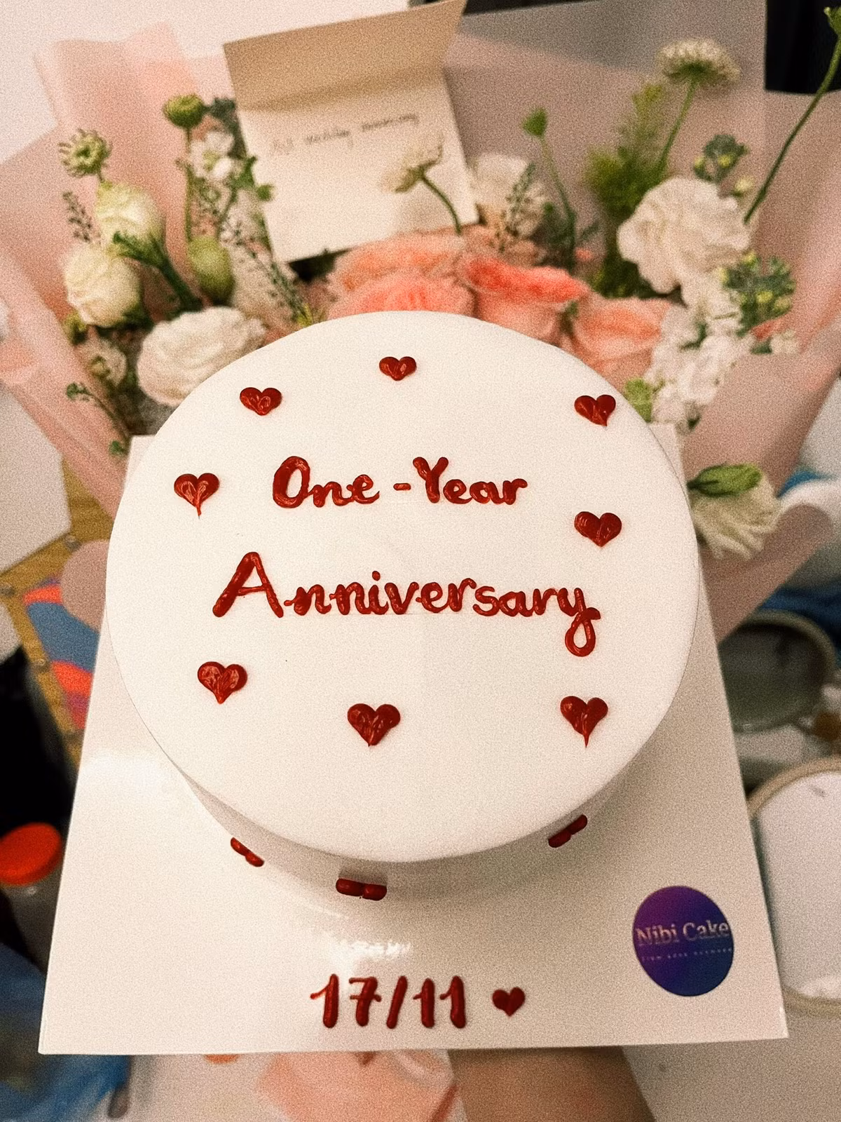 Không thể thiếu là chiếc bánh kem màu trắng tối giản, được trang trí bằng chữ "One-Year Anniversary" và ngày cưới "17/11" cùng những trái tim nhỏ màu đỏ, đánh dấu cột mốc một năm đầy ý nghĩa.