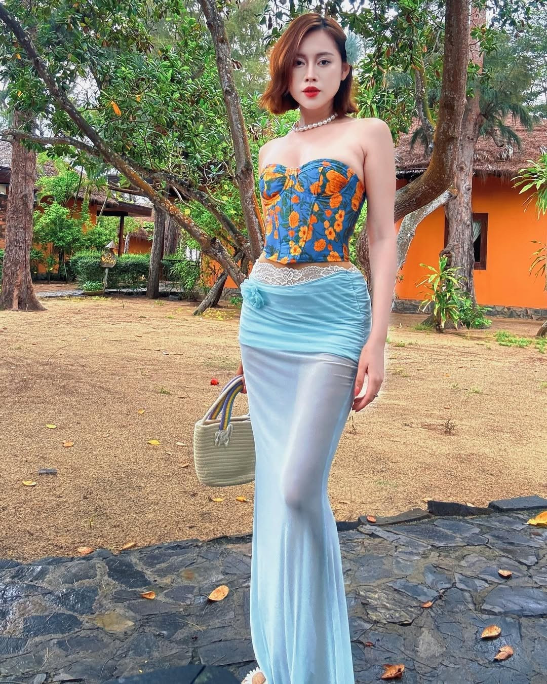 Việt Phương Thoa cũng tinh tế khi chọn thêm chuỗi vòng cổ ngọc trai, bông tai nhỏ và túi cói cầm tay, tạo nên sự hài hòa giữa hiện đại và truyền thống. Mái tóc bob được uốn nhẹ kết hợp hoa cài đầu giúp cô trông vừa ngọt ngào vừa nổi bật giữa khung cảnh đậm chất miền quê Việt.