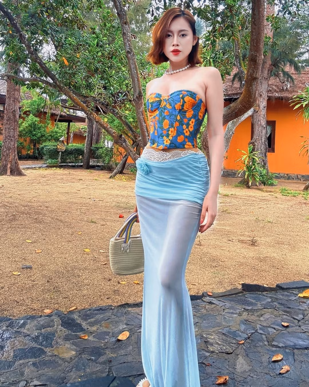 Việt Phương Thoa cũng tinh tế khi chọn thêm chuỗi vòng cổ ngọc trai, bông tai nhỏ và túi cói cầm tay, tạo nên sự hài hòa giữa hiện đại và truyền thống. Mái tóc bob được uốn nhẹ kết hợp hoa cài đầu giúp cô trông vừa ngọt ngào vừa nổi bật giữa khung cảnh đậm chất miền quê Việt.