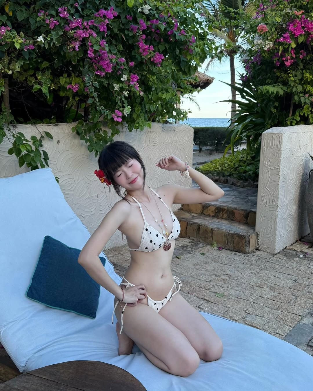 iểm nhấn là bông hoa đỏ rực cài tóc, tạo nên vẻ đẹp vừa gợi cảm vừa đáng yêu, đậm chất “tropical girl” giữa không gian biển xanh – nắng vàng.