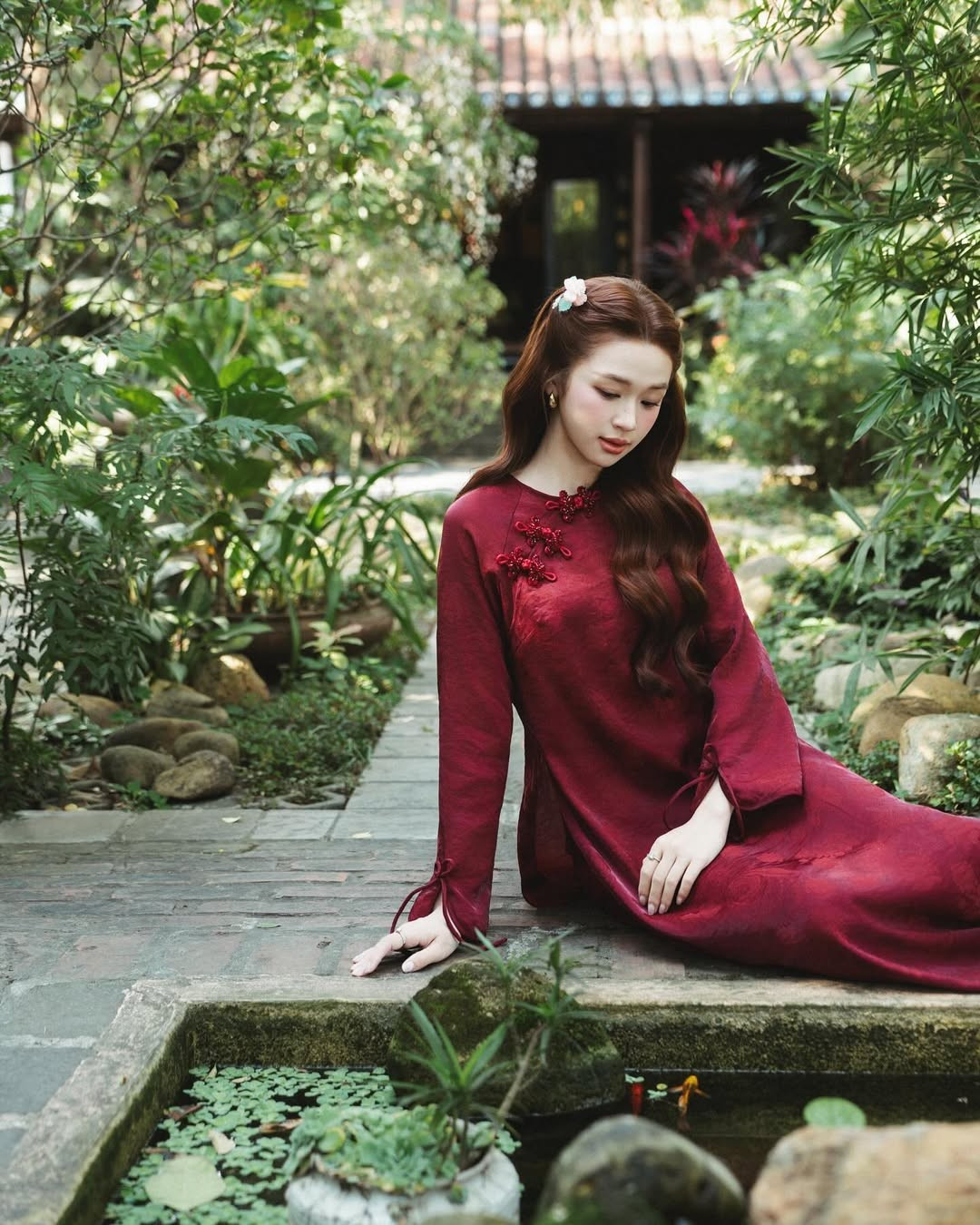 Thiết kế áo dài cổ tròn với hàng khuy tết thủ công mang đậm phong vị truyền thống. Sắc đỏ không chỉ tôn da mà còn mang ý nghĩa may mắn, giúp Karty Chang tỏa sáng rực rỡ giữa không gian xanh mướt của khu vườn. (Ảnh: IGNV)