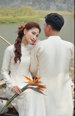 MC Mù Tạt diện chiếc áo dài trắng với họa tiết chìm tinh tế, mái tóc uốn xoăn nhẹ nhàng điểm xuyết thêm nhành hoa Thiên Điểu rực rỡ trên tay. Trong khi đó, "Hoàng tử Ả Rập" Đức Huy lịch lãm nhưng không kém phần phong độ trong thiết kế áo dài nam đồng điệu. (Ảnh: FBNV)