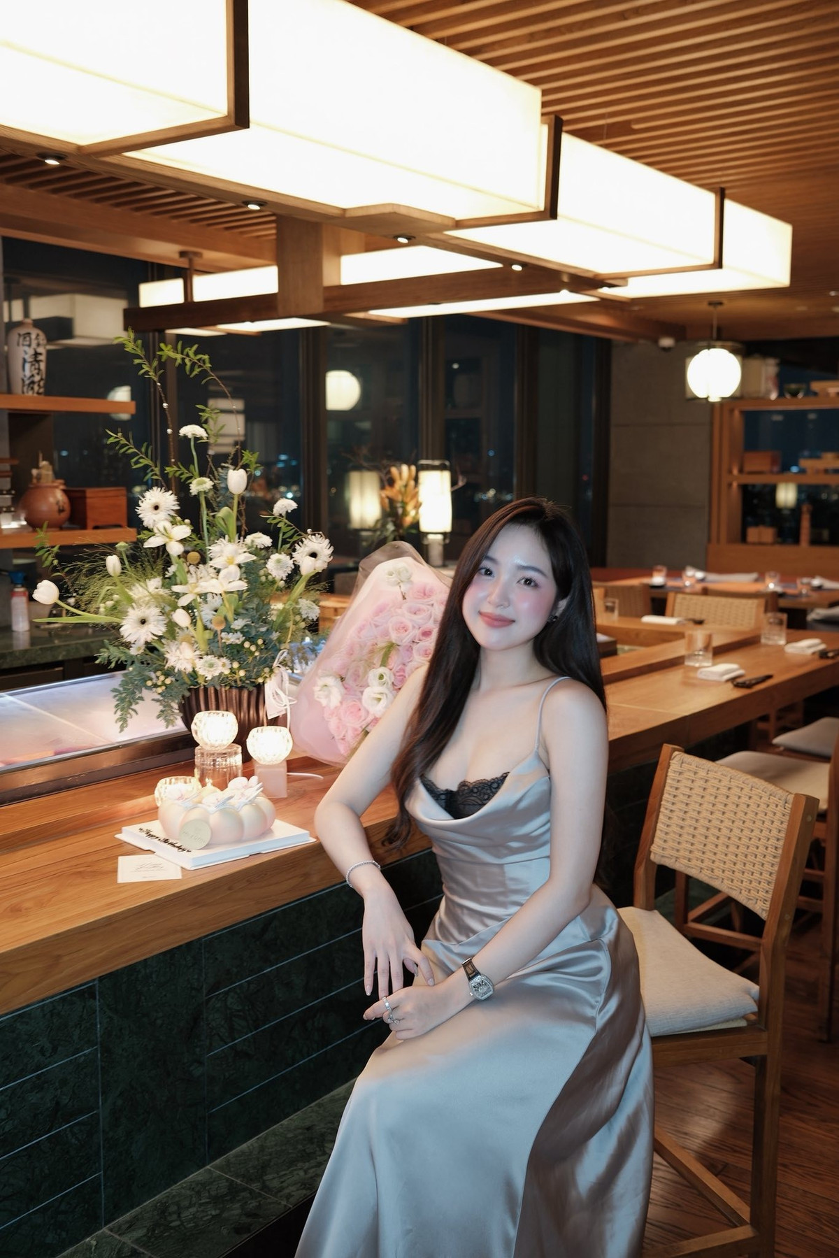 Cô tạo dáng bên bó hoa tươi lớn, chiếc bánh kem xinh xắn, hòa trong không gian nhà hàng cao cấp với quầy bar gỗ ấm cúng, ánh đèn vàng dịu. (Ảnh: FBNV)