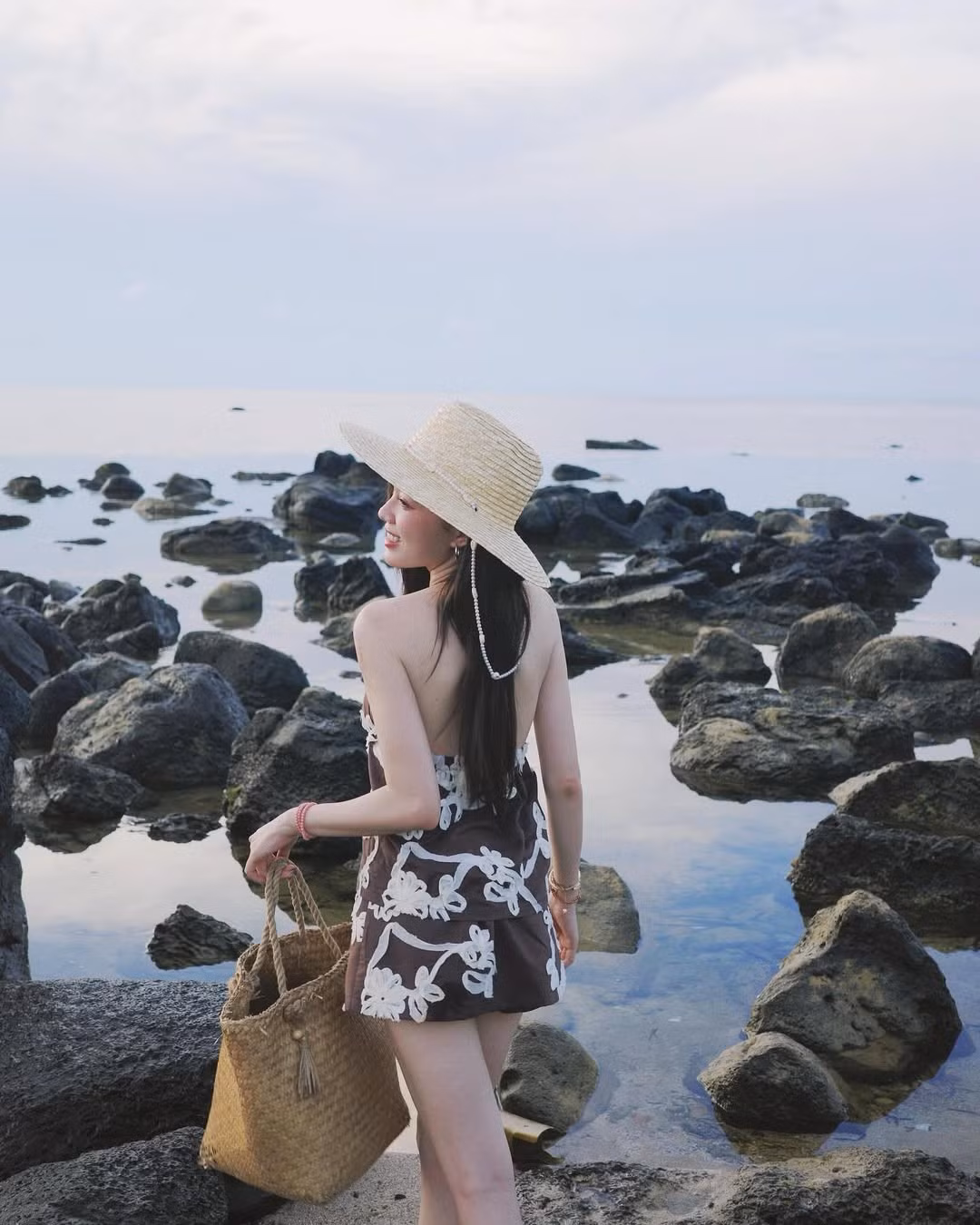 Chiếc mũ cói rộng vành và túi đan thủ công tiếp tục là hai phụ kiện chủ đạo mà Mèo Sao Hỏa ưu ái, tạo nên vẻ đẹp mộc mạc, gần gũi với thiên nhiên – rất phù hợp với khung cảnh biển trời thanh bình và đá đen hoang sơ phía sau. Bên cạnh đó, vòng cổ ngọc trai và dây chuyền vỏ sò góp phần hoàn thiện một phong cách "nàng thơ biển cả" đầy lãng mạn và thơ mộng.