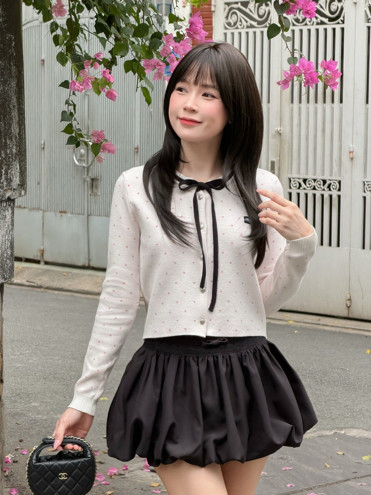 Chiếc áo khoác cardigan mỏng màu trắng kem với các điểm xuyết chấm hồng nhỏ tạo cảm giác nhẹ nhàng. Điểm nhấn là phần nơ thắt đen ở cổ và viền đen tương phản, mang lại vẻ ngoài vừa thanh lịch lại vừa có chút tinh nghịch của các nữ sinh. (Ảnh: IGNV)