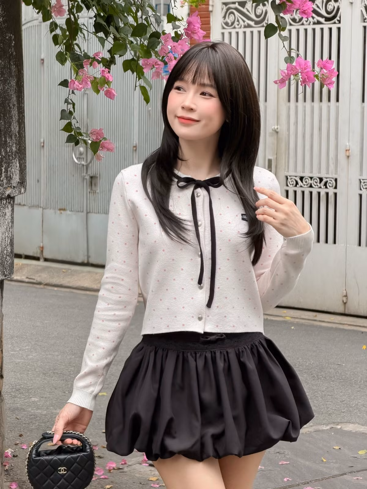 Chiếc áo khoác cardigan mỏng màu trắng kem với các điểm xuyết chấm hồng nhỏ tạo cảm giác nhẹ nhàng. Điểm nhấn là phần nơ thắt đen ở cổ và viền đen tương phản, mang lại vẻ ngoài vừa thanh lịch lại vừa có chút tinh nghịch của các nữ sinh. (Ảnh: IGNV)