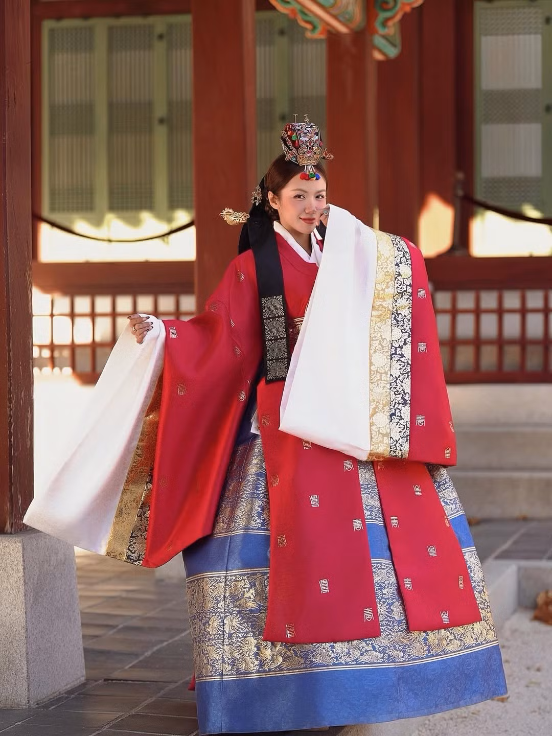 Chiếc áo khoác ngoài sử dụng tông đỏ son đậm rực rỡ, tượng trưng cho sự may mắn và quyền lực, được thêu họa tiết vàng kim tinh xảo. Áo ngoài được phối hài hòa với sắc trắng tinh khôi của áo trong và xanh kết hợp hoa văn vàng của chân váy, tạo nên tổng thể vừa truyền thống vừa nổi bật. (Ảnh: IGNV)