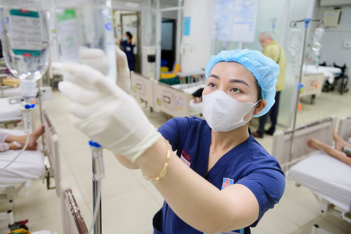 Điều dưỡng tập trung điều chỉnh chai dịch truyền cho bệnh nhân trong ca trực cận Tết. Phía sau, các giường bệnh gần như kín chỗ, nhân viên y tế liên tục di chuyển giữa các buồng. Nhịp làm việc khẩn trương, mỗi thao tác đều đòi hỏi sự chính xác và phối hợp chặt chẽ. Dịp cuối năm, lượng bệnh nhân tăng khiến áp lực tại khoa càng thêm nặng nề.