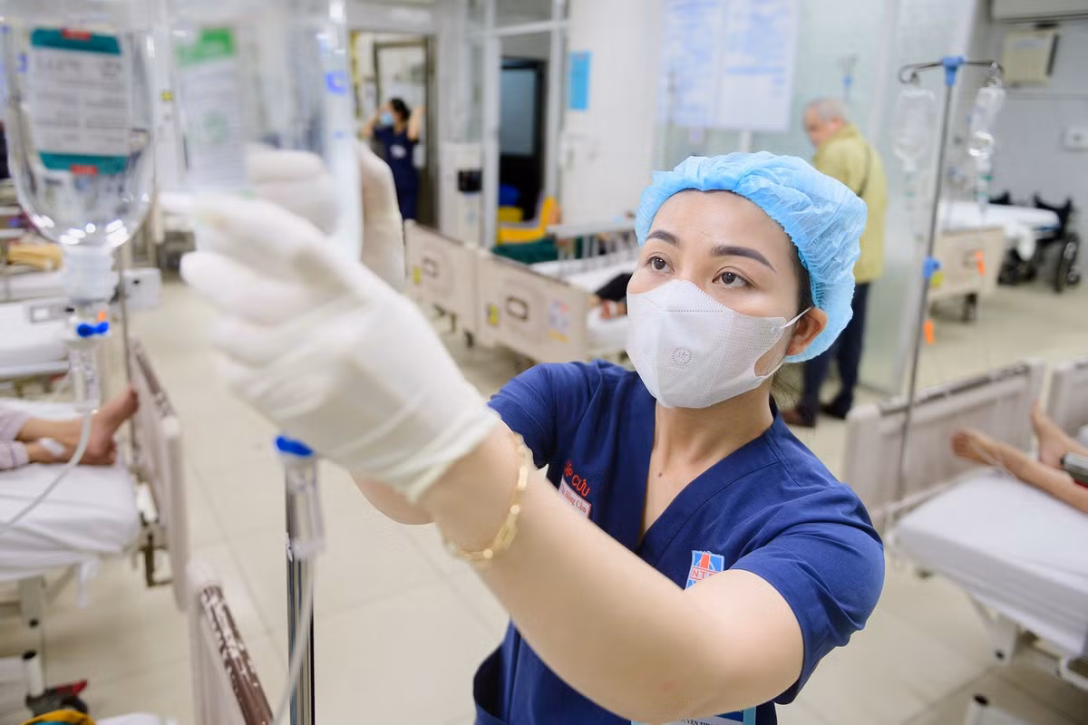 Điều dưỡng tập trung điều chỉnh chai dịch truyền cho bệnh nhân trong ca trực cận Tết. Phía sau, các giường bệnh gần như kín chỗ, nhân viên y tế liên tục di chuyển giữa các buồng. Nhịp làm việc khẩn trương, mỗi thao tác đều đòi hỏi sự chính xác và phối hợp chặt chẽ. Dịp cuối năm, lượng bệnh nhân tăng khiến áp lực tại khoa càng thêm nặng nề.
