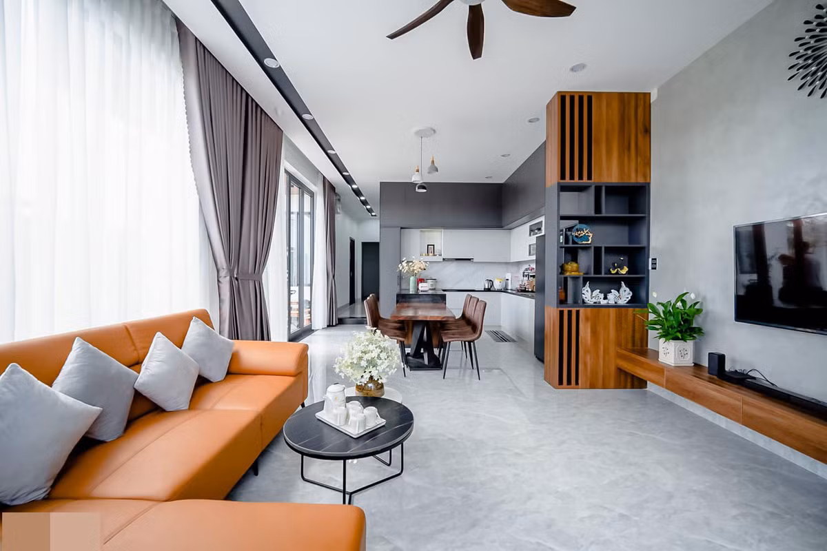 Phòng khách của ngôi nhà có nhiều khoảng sáng nhờ tường kính và cửa kính lớn. Nội thất hiện đại kiểu Hàn Quốc. Sàn lát gạch bông sáng màu dễ vệ sinh và rất mát mẻ, phù hợp thời tiết nắng nóng ở Quảng Nam. Bên cạnh phòng khách là bàn ăn và bếp nấu.