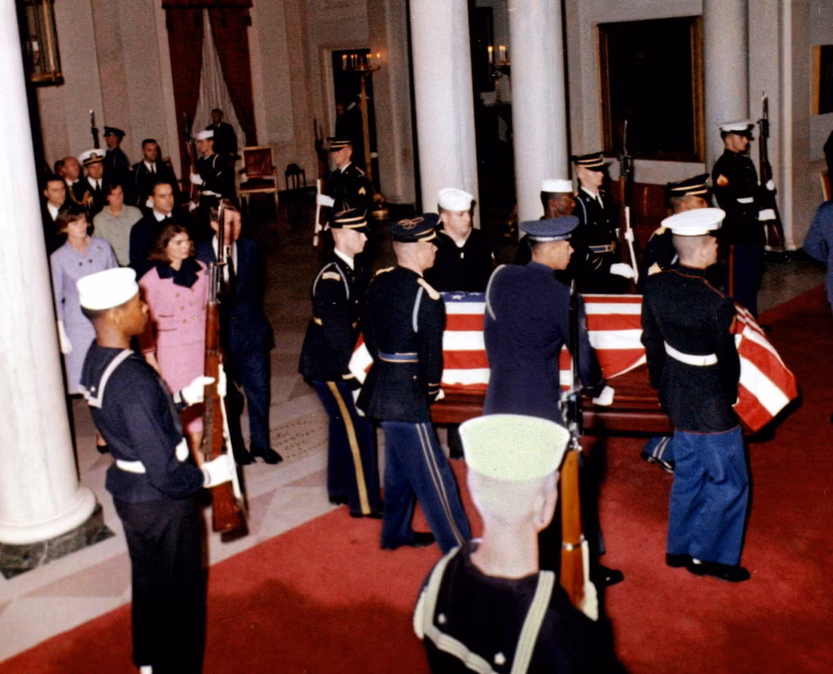 Đệ nhất phu nhân Jacqueline Bouvier Kennedy vàBộ trưởng Tư pháp Mỹ Robert F. Kennedy, anh trai ông John F. Kennedy, bước vào tiền sảnh của Nhà Trắng sau quan tài của tổng thống. Thi hài Tổng thống Kennedy được chuyển từ Dallas về Washington vào sáng sớm ngày 23/11/1963.