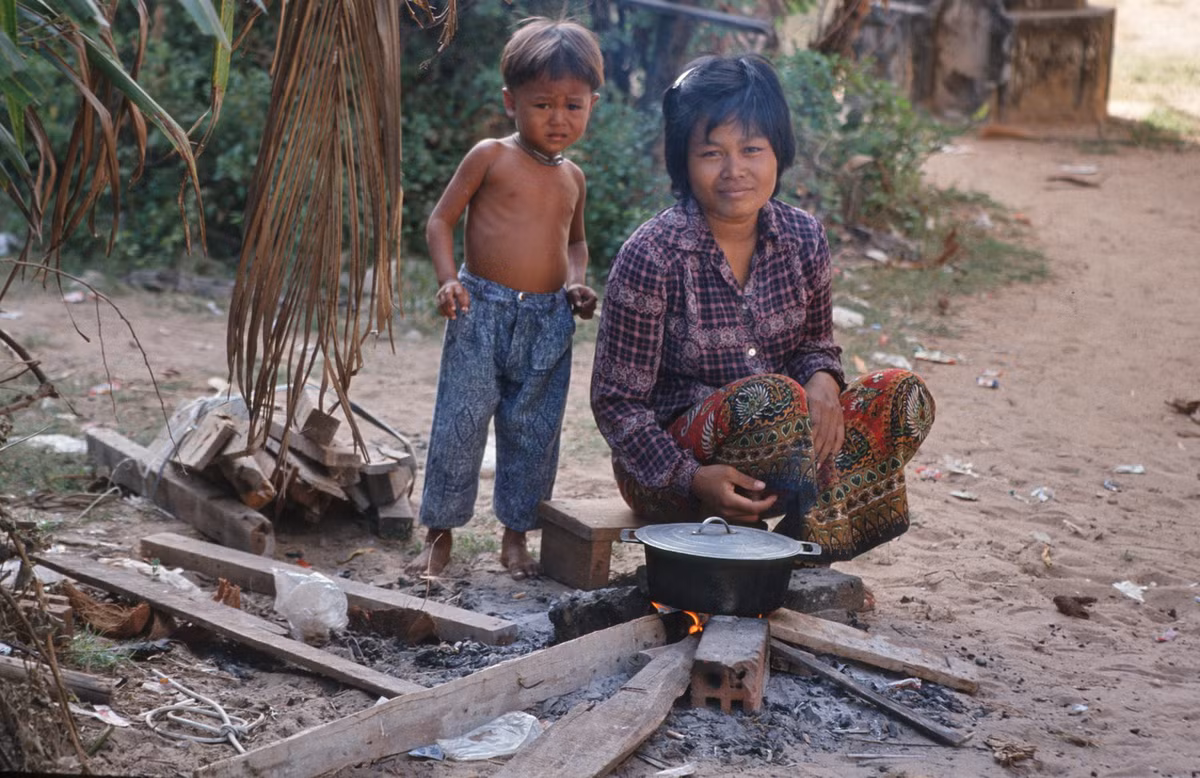 Bà mẹ và đứa con chuẩn bị bữa ăn bằng bếp củi thô sơ ở ngoài trời.