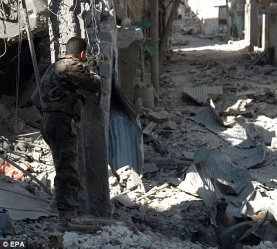 Một người lính quân đội chính phủ Assad đứng nấp đằng sau một khối bê tông xung quanh là đống đổ nát, hoang tàn.