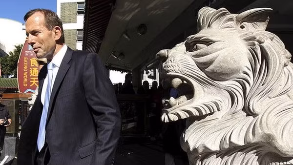 Ông Abbott cạnh một con sư tử đá, phía xa là tấm bảng hiệu có chữ Hoa và tiếng Anh mang nghĩa "Nuốt cả thế giới".
