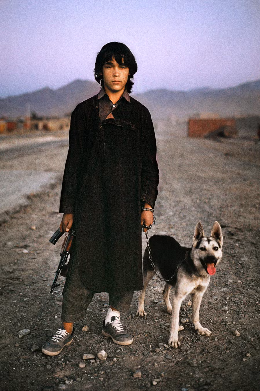 Cậu bé người Afghanistan dắt chó, tay cầm súng trường, đứng bên con đường dẫn đến Kandahar.