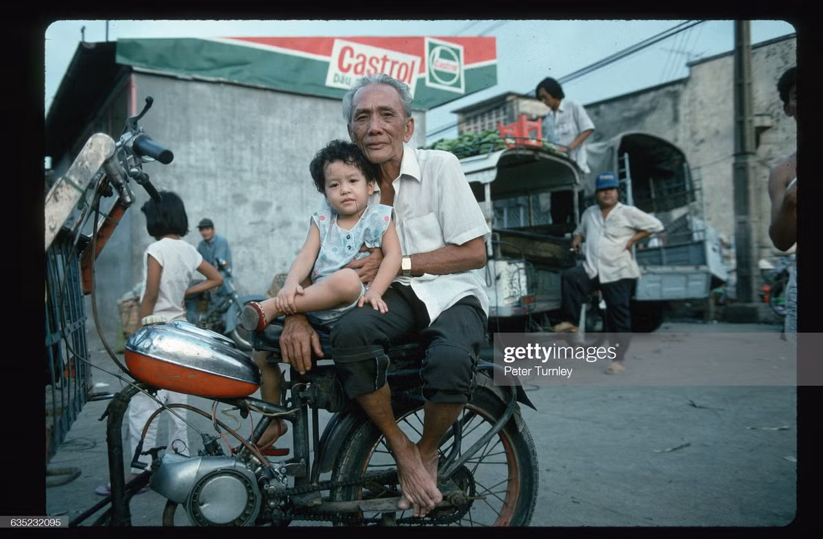 Hai ông cháu ngồi trên chiếc xe máy cạnh cửa hàng xăng dầu Castrol.