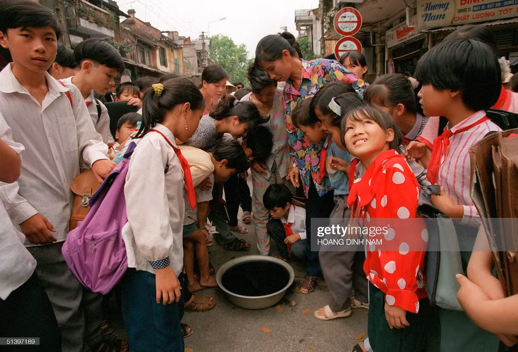 Trẻ em và người lớn hào hứng vây quanh một cái chậu chứa nước pha mực để quan sát hình ảnh phản chiếu của nhật thực ngày 24/10/1995.