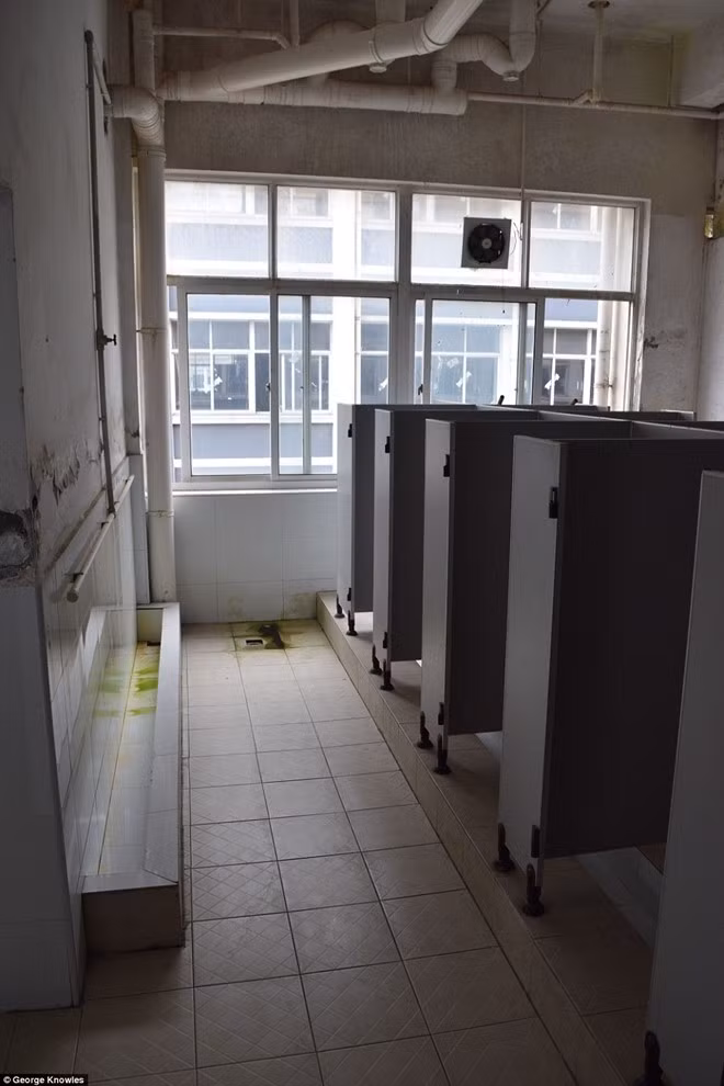 Phòng vệ sinh bẩn thỉu với bệ xí được thiết kế ngồi xổm, tường tróc ra thành từng mảng.