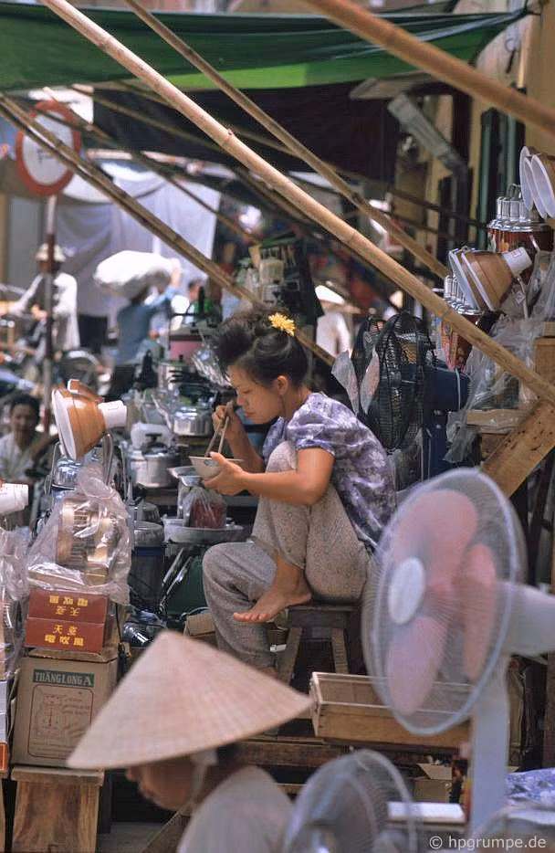 Người bánh hàng ngồi ăn trưa giữa đống hàng hóa gồm quạt điện, đèn bàn, nồi áp suất, phích nước...