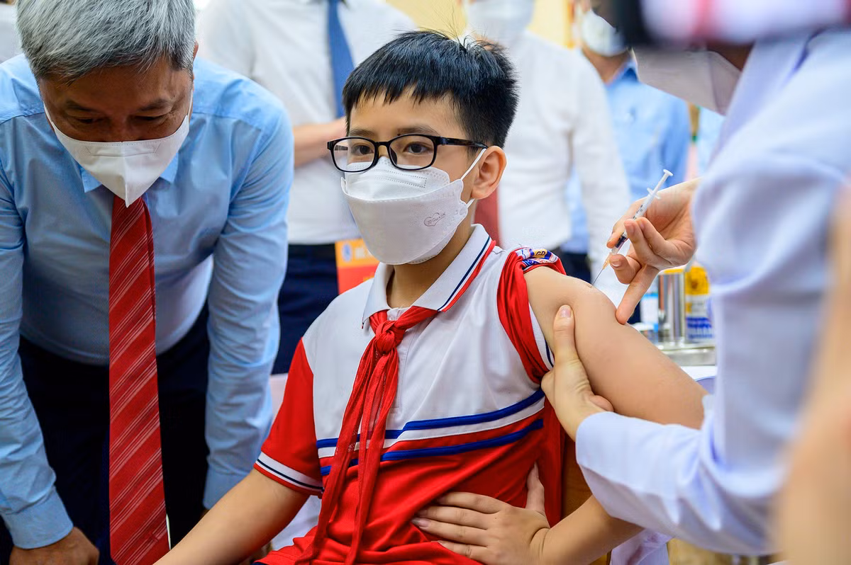 Tuấn Minh (học sinh lớp 6A2) là người đầu tiên được tiêm vắc xin. "Em không sợ tiêm nên khá bình tĩnh. Tiêm chủng tốt cho cả sức khỏe bản thân và cộng đồng", Minh chia sẻ. Thứ trưởng Bộ Y tế Nguyễn Trường Sơn (bên trái) đến trực tiếp phòng tiêm để nói chuyện, động viên học sinh cùng cán bộ y tế tham gia chiến dịch.