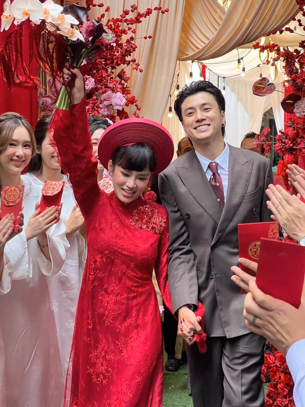 Trong ngày trọng đại, Ngọc Mai diện áo dài đỏ, đội mấn truyền thống, cầm bó hoa lan trắng. Vương Anh Ole chọn bộ suit màu xám lịch lãm, nắm tay cô dâu bước qua lối đi phủ kín hoa đỏ và hồng.